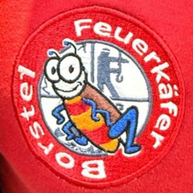 Aufnäher auf einem roten Kleidungsstück: In der Mitte ist ein Käfer, hinter ihm ist das Feuerwehr-Emblem, das die vier Hauptaufgaben der Feuerwehr darstellt: Retten / Löschen / Bergen / Schützen. Drumherum steht kreisrund: "Feuerkäfer Borstel".