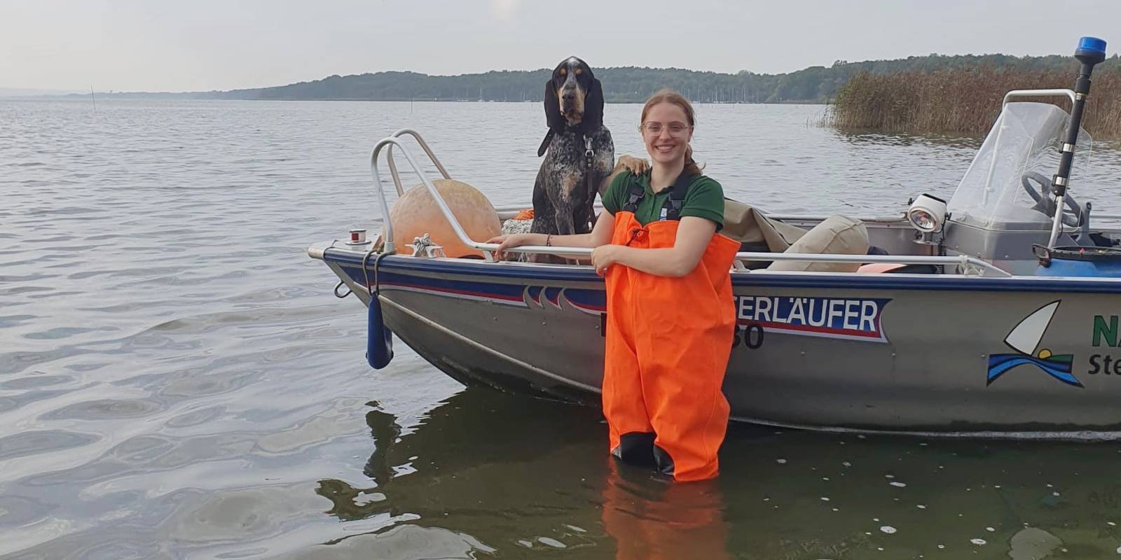 Eine Frau trägt eine orangefarbene Wathose und steht vor einem Boot im Wasser des Steinhuder Meeres. Im Boot sitzt ein Hund und schaut mit der Frau in die Kamera.
