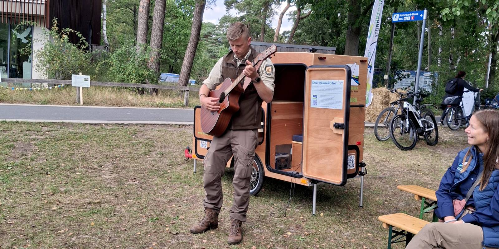 Ein Mann trägt Outdoor-Kleidung, steht vor einem besonderen Fahrradanhänger aus Holz mit Mikrofontechnik und Infomaterial, er spielt auf einer Gitarre.