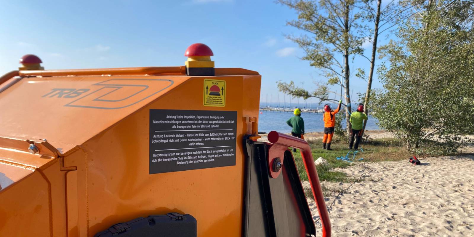 Blick über einen mobilen Häcksler zu Personen, die in der Ferne Bäume begutachten.