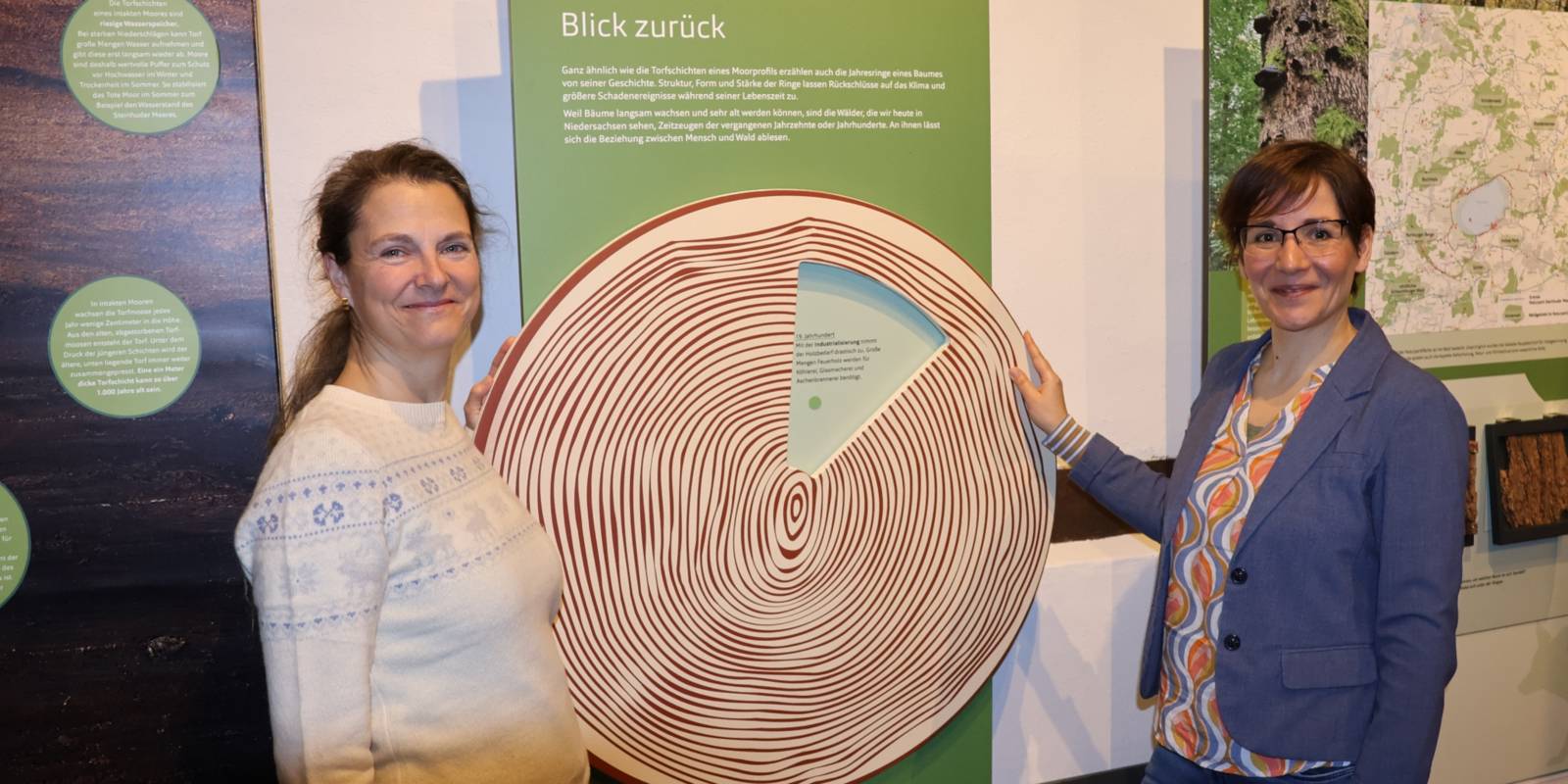 Zwei Frauen stehen in einem Ausstellungsbereich mit Mitmach-Aktionen an einer Drehscheibe in Baumscheibenoptik mit Jahresringen. Ein Sichtfenster in der Drehscheibe lässt immer nur einen kleinen Ausschnitt des darunter liegenden Textbereiches frei. An der Wand darüber steht unter anderem: "Blick zurück"