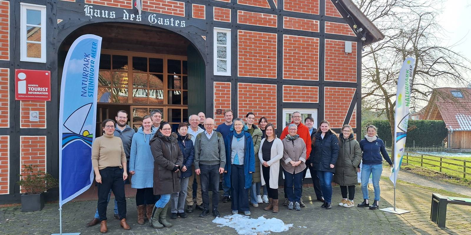 Zwanzig Personen stehen zwischen zwei Strandflaggen vor einem Fachwerkhaus.
