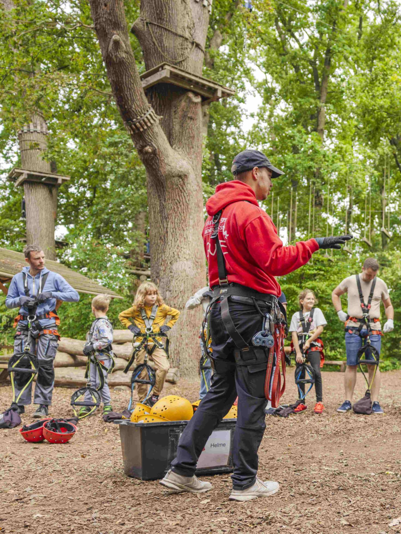 Eine Person steht neben einer Kiste mit Helmen und leitet Personen an, die sich Klettergeschirre anlegen