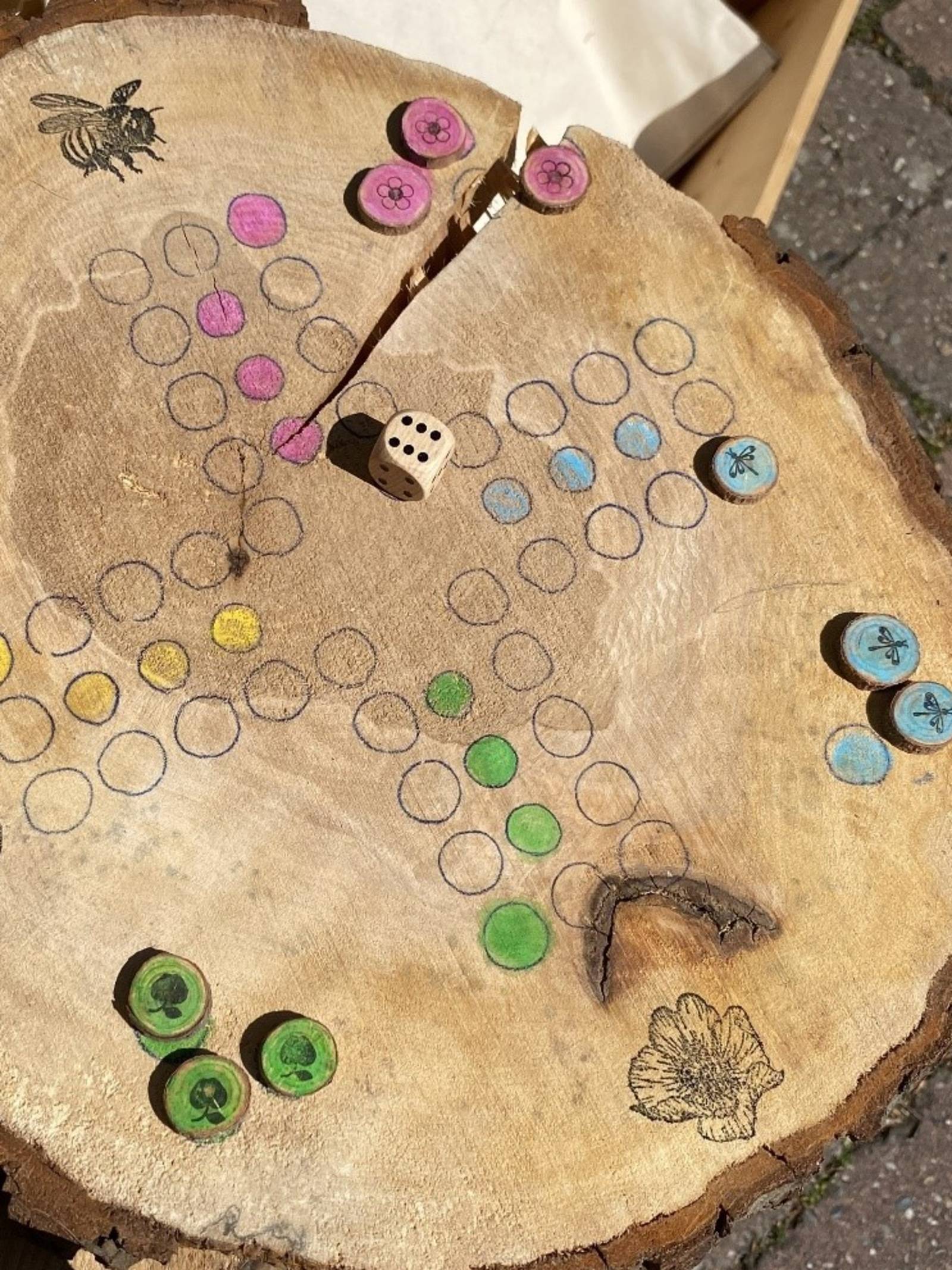 Auf eine Baumscheibe ist das Spielbrett eines Würfelspiels gezeichnet. Felder sind farbig markiert und Stempel verzieren das Spielbrett.