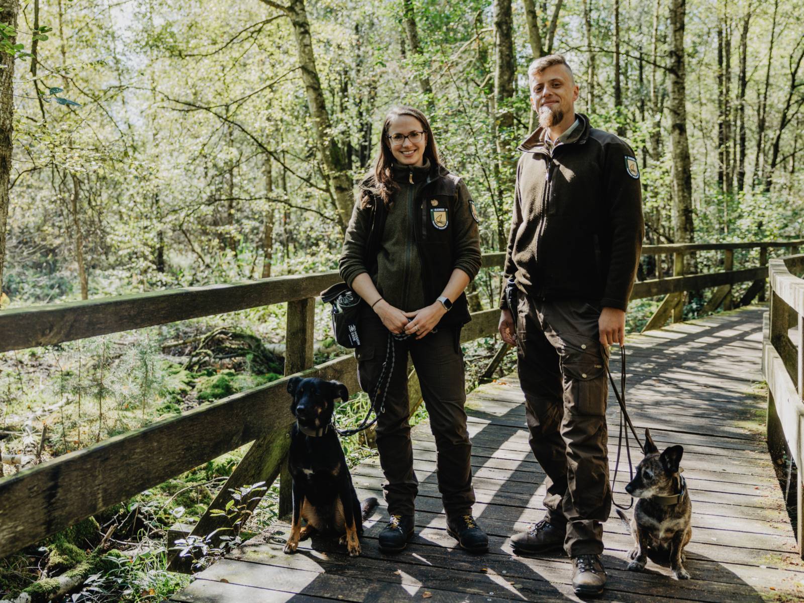 Eine Rangerin und ein Ranger stehen nebeneinander auf einem Holzsteg, zu ihren Füßen sitzen zwei Hunde, die Menschen halten die Hundeleinen.