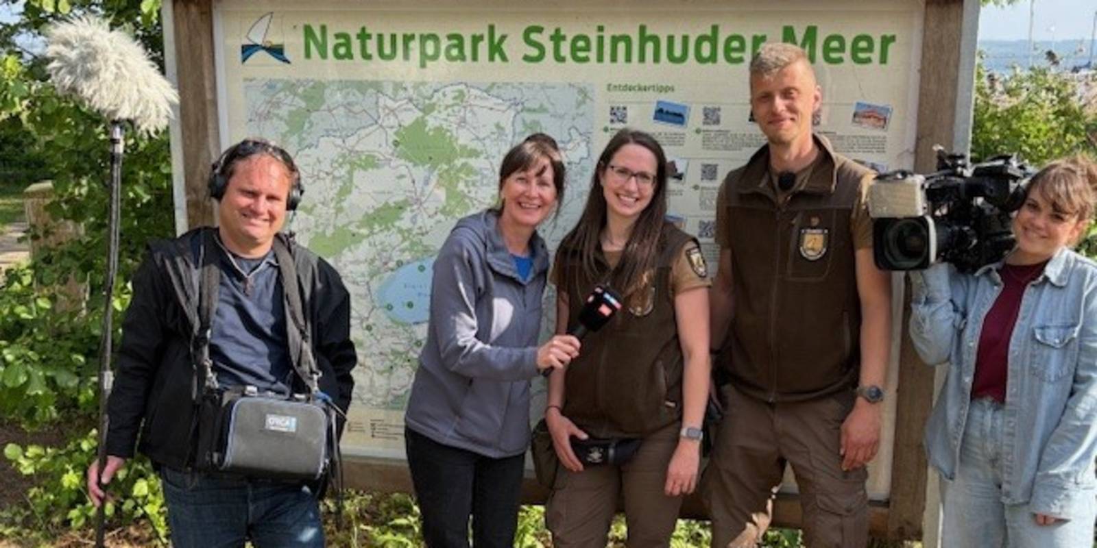 5 Personen stehen vor einer Infotafel im Naturpark Steinhuder Meer, zu sehen sind auch zwei Mikrofone und eine Kamera eines Fernseh-Teams.