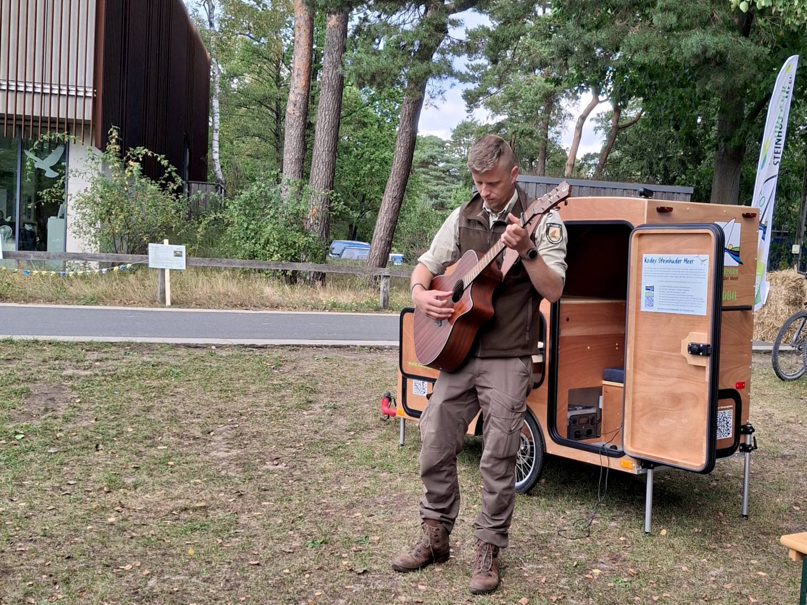 Ein Mann trägt Outdoor-Kleidung, steht vor einem besonderen Fahrradanhänger aus Holz mit Mikrofontechnik und Infomaterial, er spielt auf einer Gitarre.