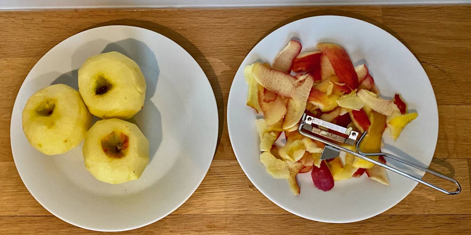 Zwei Teller stehen auf einem Tisch. Auf dem linken Teller liegen drei geschälte Äpfel, auf dem rechten Teller liegen Apfelschalen und ein Schäler.