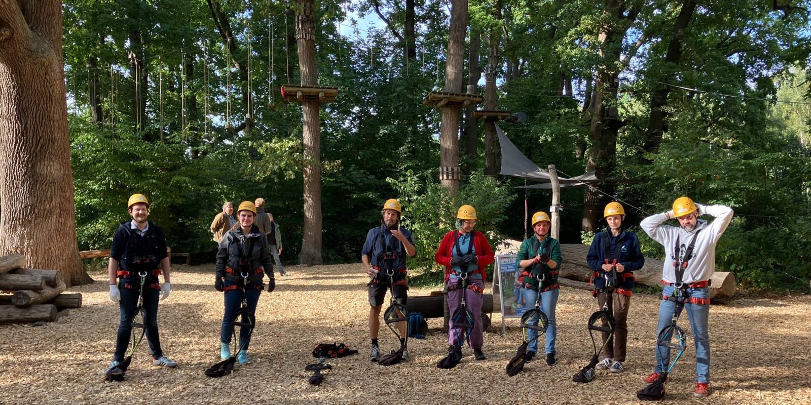 Sieben Personen stehen nebeneinander und tragen Kletterausrüstung. Im Hintergrund sind weitere Personen und ein Kletterpark im Wald.