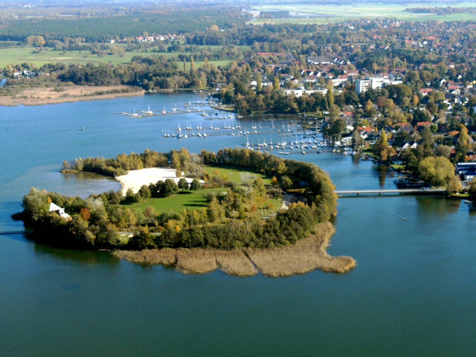 Die Badeinsel im Steinhuder Meer, aufgenommen aus der Luft. Die kleine Insel ist mit Gras und Bäumen bewachsen, es gibt eine kleine Bucht ist mit hellem, weißen Sandstrand. Ein Steg verbindet die Badeinsel mit dem Festland.