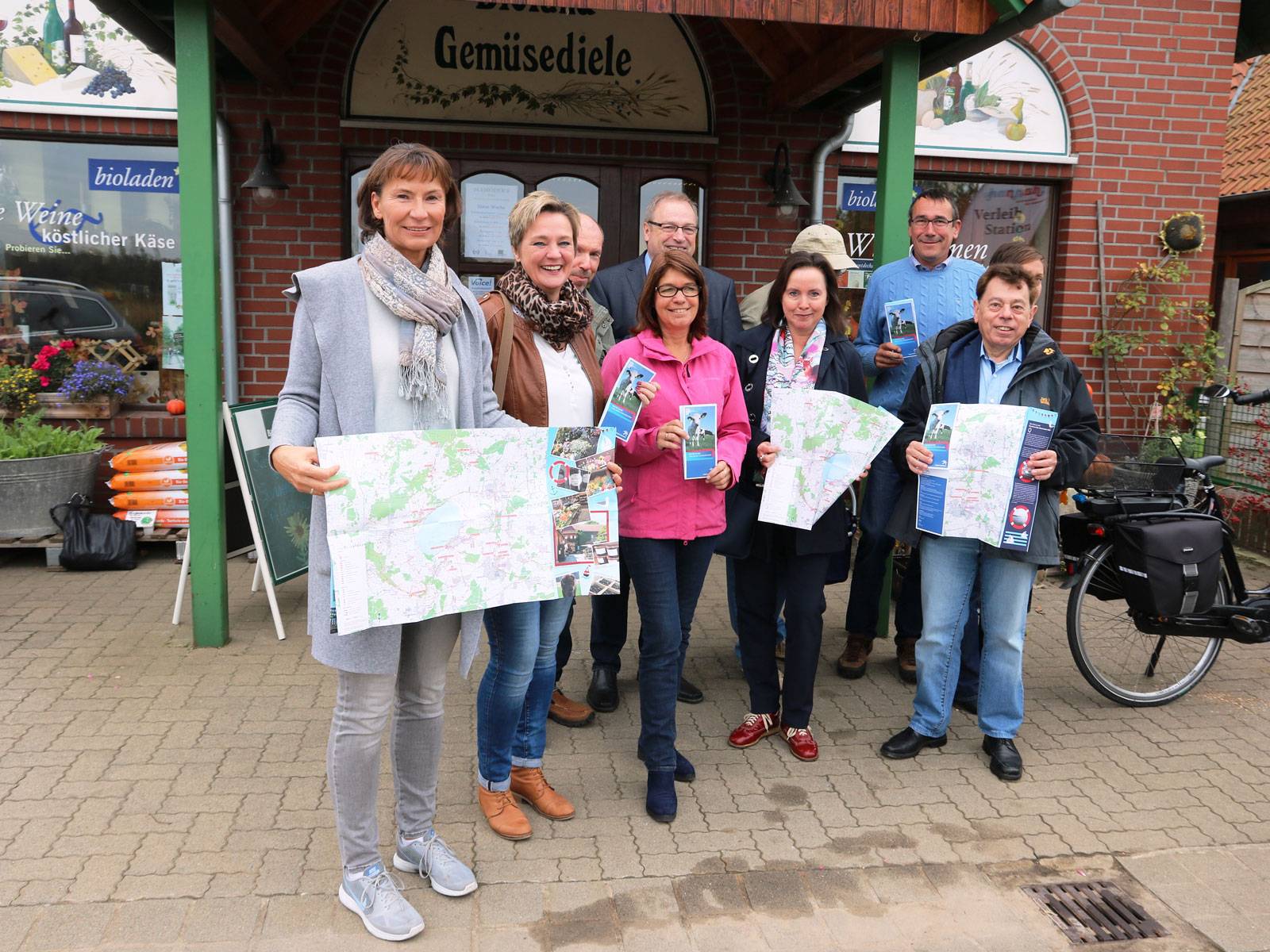 Eine kleine Gruppe Männer und Frauen, die eine Broschüre in den Händen halten, vor einem Laden in einem Backsteingebäude. 