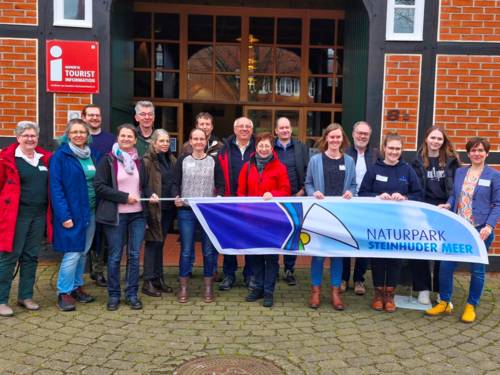 16 Personen haben sich vor einem Fachwerkhaus für ein Gruppenfoto aufgestellt. Die Personen halten gemeinsam eine Strandflagge mit dem Logo Naturpark Steinhuder Meer.