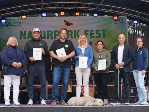 Sieben Personen stehen auf einer Bühne, vier der Personen halten eine Urkunde in den Händen. Mittig zu deren Füßen liegt ein Hund.