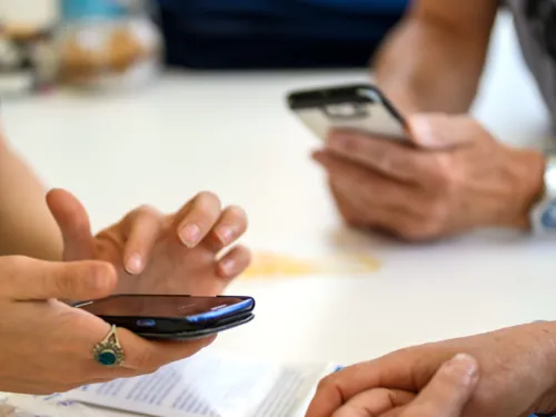 Zwei Personen sitzen an einem Tisch und machen etwas mit Smartphones. Eine dritte Person hat die leeren Hände vor sich auf dem Tisch ineinander verschränkt.