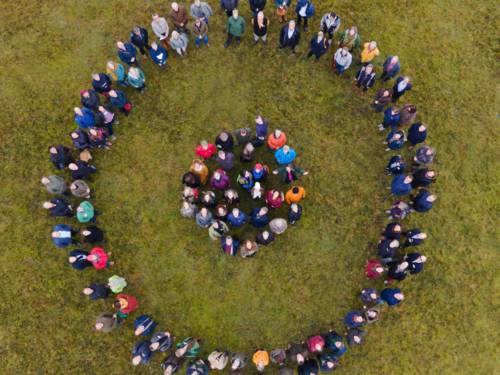 Foto aus der Vogelperspektive: Männer und Frauen haben sich auf einer Rasenfläche so aufgestellt, dass ein äußerer Kreis einen inneren Kreis umschließt. So bilden Sie aus Menschen das Logo der Nationalen Naturlandschaften.