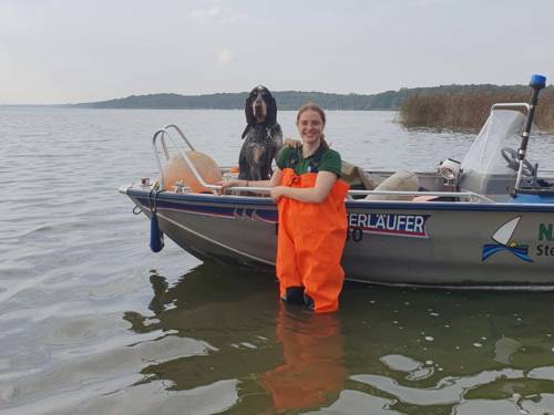 Eine Frau trägt eine orangefarbene Wathose und steht vor einem Boot im Wasser des Steinhuder Meeres. Im Boot sitzt ein Hund und schaut mit der Frau in die Kamera.