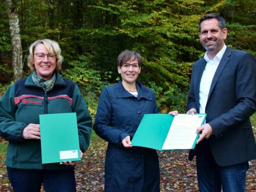 Zwei Frauen und ein Mann halten Mappen mit Dokumenten und schauen in die Kamera.