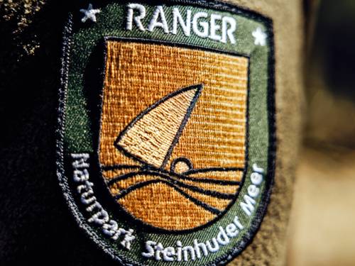 Aufnäher auf dem Ärmel einer Uniform eines Rangers im Naturpark Steinhuder Meer: "Ranger Naturpark Steinhuder Meer", dazu das Logo des Naturparks Steinhuder Meer, einem Segelboot mit Wasser und Land.