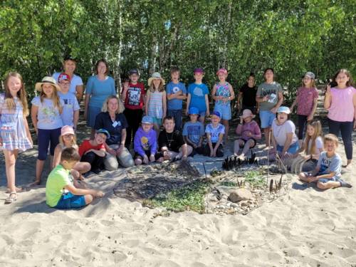 23 Kinder und drei Frauen hocken oder stehen im Halbkreis um ein Mandala, das im Sand aus Naturmaterialien geschaffen wurde.