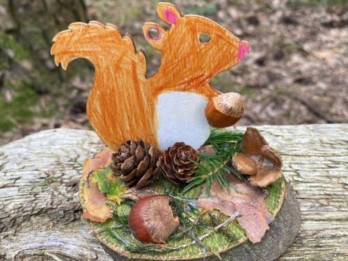 Ein gebasteltes Eichhörnchen steht auf einer Baumscheibe, die Halt gebende Baumscheibe ist mit Fundstücken aus dem Wald dekoriert, zum Beispiel mit Tannenzapfen, Kastanien und Moos.