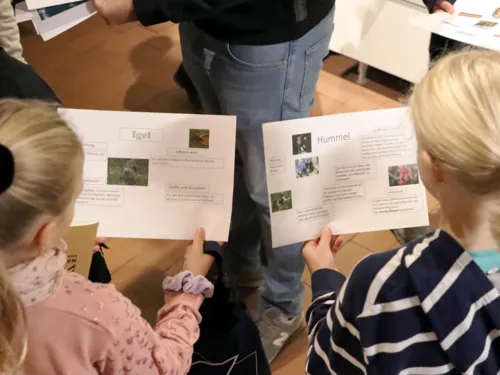 Zwei M&auml;dchen halten jeweils ein Blatt Papier mit Informationen: Links &uuml;ber den Igel, rechts &uuml;ber die Hummel.