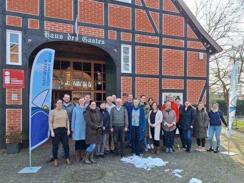 Zwanzig Personen stehen zwischen zwei Strandflaggen vor einem Fachwerkhaus.