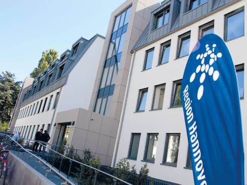 Ein modernes Gebäude mit einer Rampe für einen barrierefreien Zugang. Im Eingangsbereich stehen zwei Personen und eine weitere Person schließt ihr Fahrrad an. Vorne rechts im Bild ist ein blauer Aufsteller mit der Aufschrift "Region Hannover" zu sehen (das Wort Hannover ist zum Teil abgeschnitten).