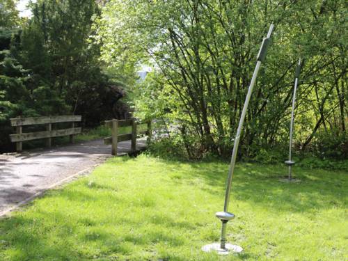 Metallstangen auf einer Rasenfläche, dahinter ein Weg und eine Brücke