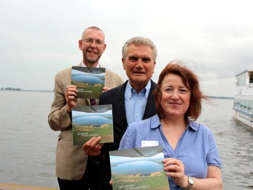 Prof. Dr. Axel Priebs (von links), Umweltdezernent der Region Hannover, Dr. Michael Arndt, Präsident des Verbandes deutscher Naturparke und Andrea Zörnig, Geschäftsführerin des Naturparks Steinhuder Meer stehen versetzt hintereinander am Ufer des Steinhuder Meeres. Jede Person hält eine Ausgabe des Buches "Naturpark Steinhuder Meer" in den Händen. Im Hintergrund sind die Festung Wilhelmstein und ein Ausflugsboot zu erkennen.