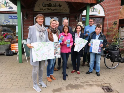 Eine kleine Gruppe M&auml;nner und Frauen, die eine Brosch&uuml;re in den H&auml;nden halten, vor einem Laden in einem Backsteingeb&auml;ude. 