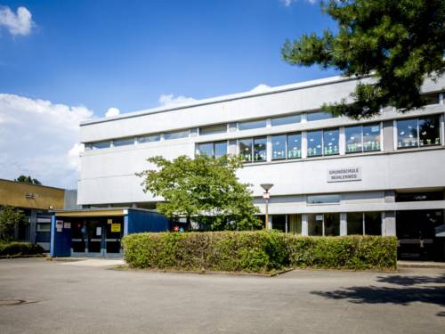 Eingang zu einem zweigeschossigen Schulgebäude, an dessen Fenster im Obergeschoss selbstgemachte Papierblumen befestigt sind. Darunter befindet sich das Schild "Grundschule Mühlenweg".