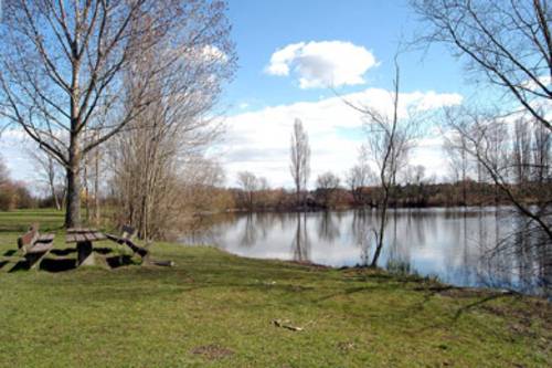 Ein See in dem sich der blaue Himmel spiegelt, im Vordergrund eine Wiese mit einem Tisch und Bänken aus Holz.