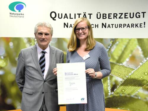 Ein Mann und eine Frau stehen nebeneinander, die Frau hält ein Zertifikat in ihren Händen. Im Hintergrund zeigt ein Banner die Blätter einer Grünpflanze, darüber steht der Text "Qualität überzeugt. Natürlich Naturparke!"