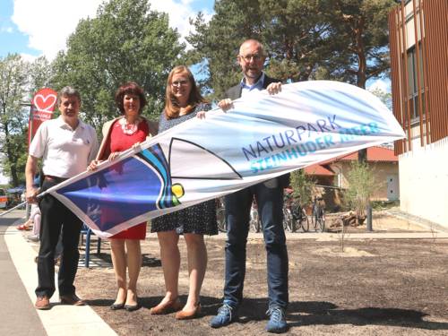 Zwei Männer und eine Frau stehen neben dem Naturparkhaus in Mardorf und halten eine Strandflagge, auf der das Logo des Naturparks Steinhuder Meer abgebildet ist.