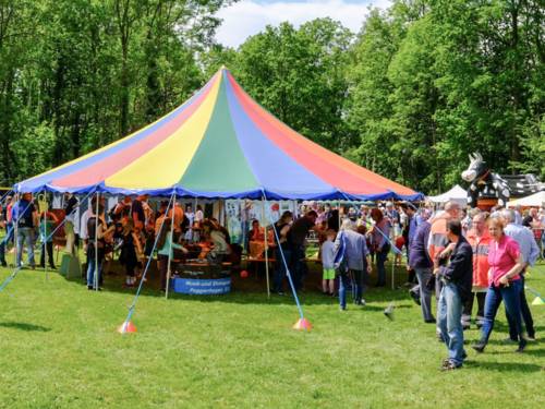 Ein Zirkuszelt steht auf einer grünen Wiese, die Seiten sind offen, so dass aus allen Richtungen Menschen hinein und heraus spazieren können. Auf dem Platz herrscht insgesamt buntes Treiben.