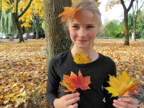 Ein Mädchen steht unter einem Baum. Herbstlaub liegt auf dem Fußboden, auch auf dem Kopf des Mädchens liegt ein Blatt und es hält Blätter in den Händen.