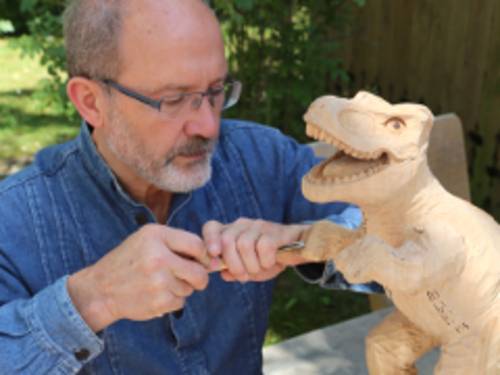 Ein Mann bearbeitet einen Holzklotz, dieser nimmt die Form eines Dinosauriers an.
