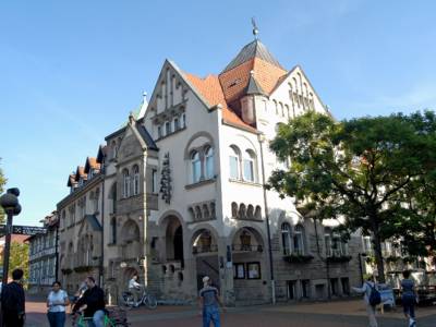 Mehrgeschossiges Gebäude mit Rundbogenfenstern, mehreren Spitzdächern und einem Glockenspiel an der Außenfassade.