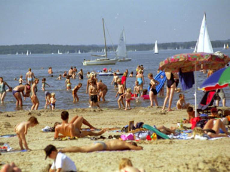im Vordergrund Sandstrand mit sich sonnenden Menschen aller Altersklassen, im Uferbereich Kinder und Erwachsene beim Spielen und Schwimmen, dahinter auf dem Wasser weisse Segelboote