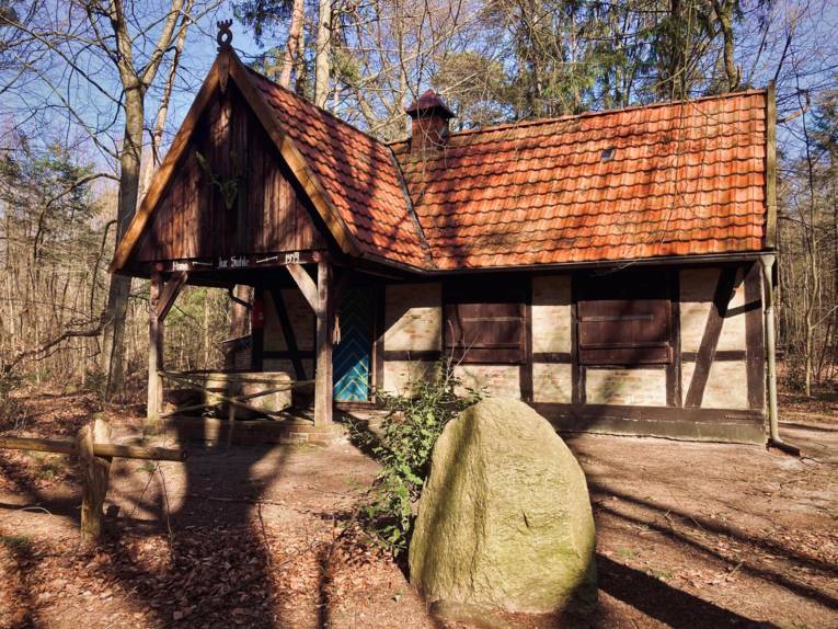 Im Hintergrund steht ein altes, kleines Dachwerkgebäude mit Vordach, davor liegt ein Findling.