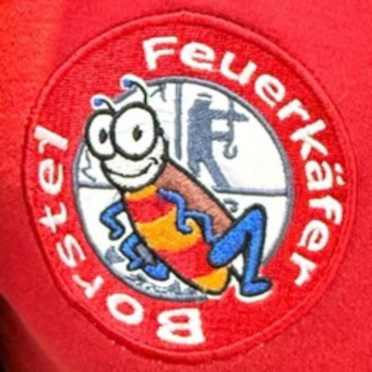 Aufnäher auf einem roten Kleidungsstück: In der Mitte ist ein Käfer, hinter ihm ist das Feuerwehr-Emblem, das die vier Hauptaufgaben der Feuerwehr darstellt: Retten / Löschen / Bergen / Schützen. Drumherum steht kreisrund: "Feuerkäfer Borstel".