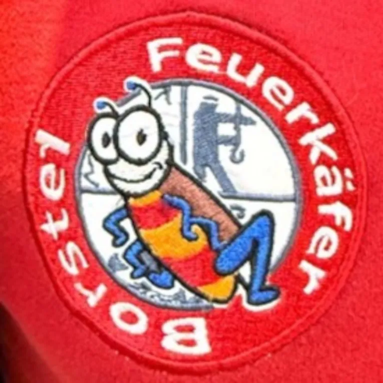 Aufn&auml;her auf einem roten Kleidungsst&uuml;ck: In der Mitte ist ein K&auml;fer, hinter ihm ist das Feuerwehr-Emblem, das die vier Hauptaufgaben der Feuerwehr darstellt: Retten / L&ouml;schen / Bergen / Sch&uuml;tzen. Drumherum steht kreisrund: "Feuerk&auml;fer Borstel".