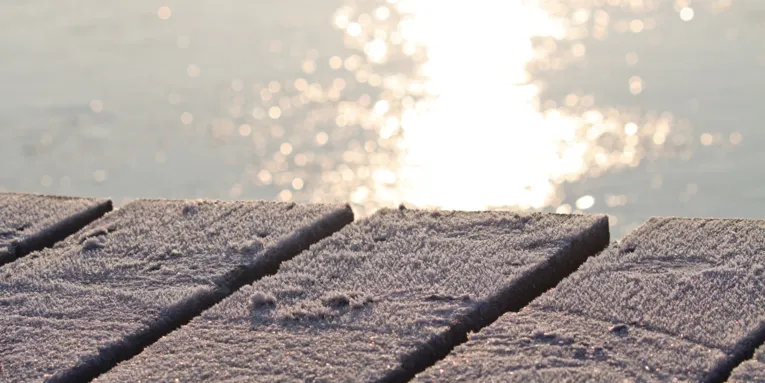 Blick &uuml;ber einen von Eis &uuml;berzogenen Steg. Hinter und unter den vereisten Holzbrettern schimmert eine von der Sonne beschienene Wasserfl&auml;che.