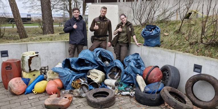 Drei Personen stehen hinter einem Haufen aus Müll und reckten jeweils einen Daumen vor der Brust nach oben.