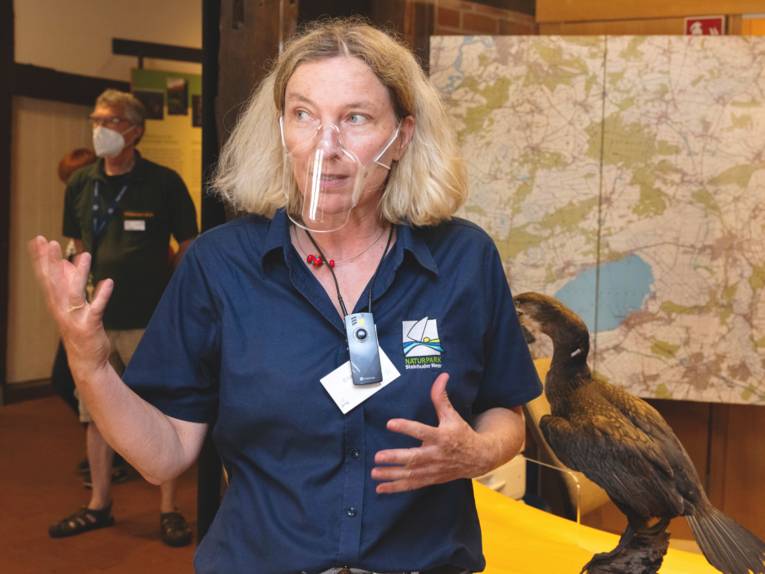 Eine Frau trägt ein Poloshirt mit dem Naturparklogo und erklärt etwas, ihre Hände sind zu einer Geste ausgestreckt.