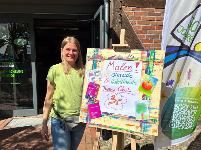 Eine Frau steht neben einer Staffelei, auf der Staffelei sind Infos zu einem Malangebot.