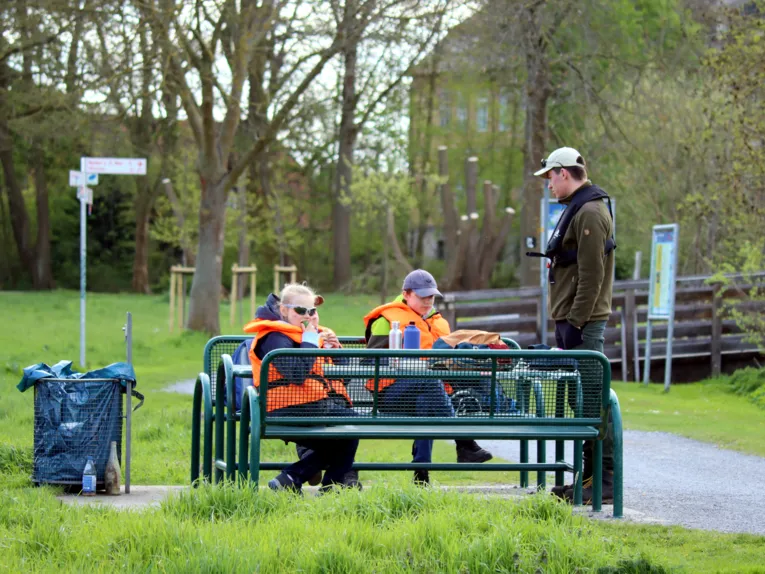 Drei Kinder sitzen auf einer Bank und verpflegen sich, daneben steht ein Naturparkmitarbeiter.