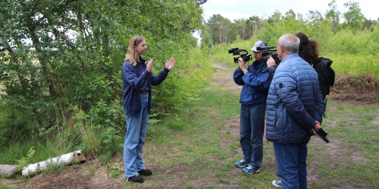 Eine Frau steht in einer Moorlandschaft. Zwei Männer halten jeweils eine Kamera auf sie, ein weiterer Mann steht daneben.