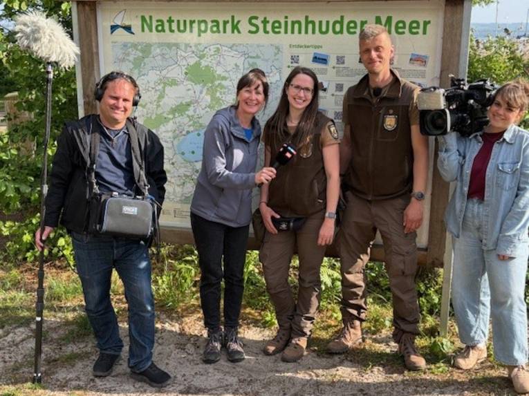 5 Personen stehen vor einer Infotafel im Naturpark Steinhuder Meer, zu sehen sind auch zwei Mikrofone und eine Kamera eines Fernseh-Teams.