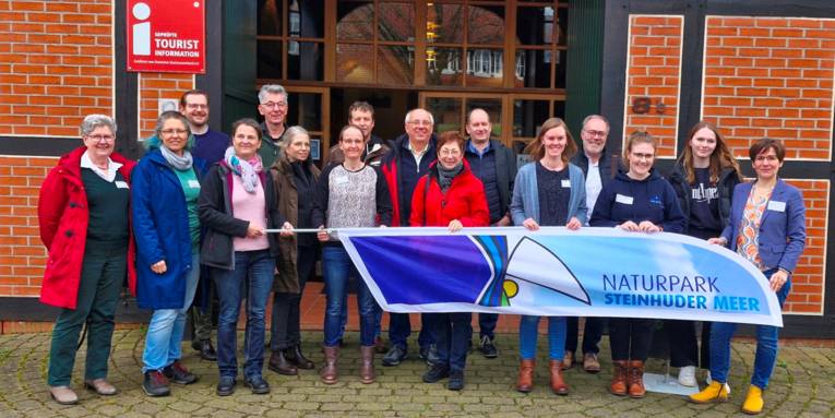 16 Personen haben sich vor einem Fachwerkhaus für ein Gruppenfoto aufgestellt. Die Personen halten gemeinsam eine Strandflagge mit dem Logo Naturpark Steinhuder Meer.